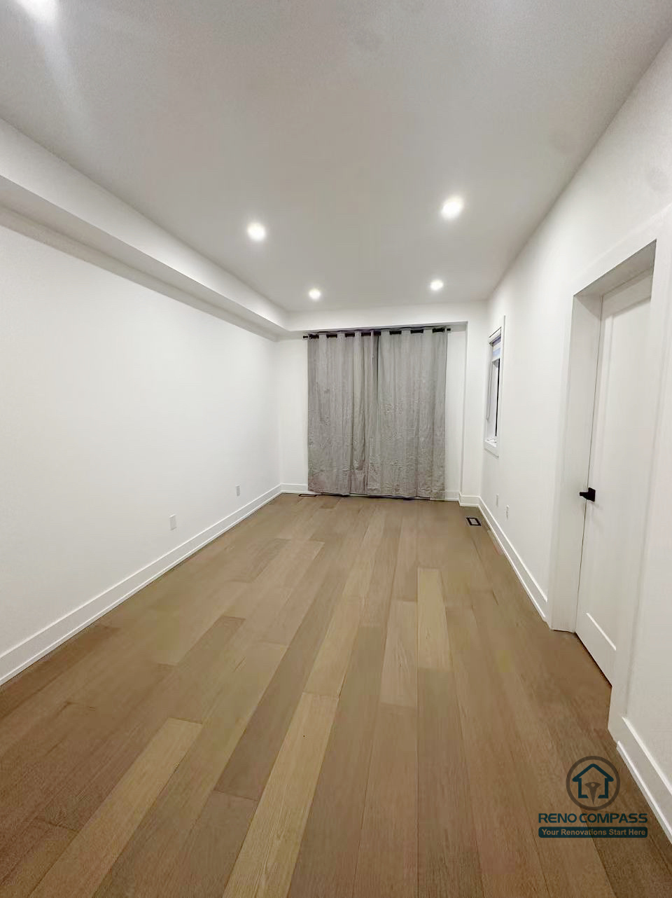 这张图片展示了 Markham 室内装修项目中的多功能长形房间空间。整体设计以简洁实用为主，通过统一的浅色墙面、木地板与嵌入式天花照明，拉伸空间纵深感，使房间在视觉上更加开阔通透。末端窗户与窗帘设计为空间引入自然采光，适合灵活用作卧室、书房或家庭活动区域。该空间由 Reno Compass 负责施工与细节处理，满足长期居住与多样化使用需求。