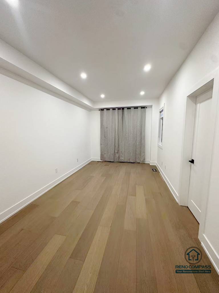 这张图片展示了 Markham 室内装修项目中的多功能长形房间空间。整体设计以简洁实用为主，通过统一的浅色墙面、木地板与嵌入式天花照明，拉伸空间纵深感，使房间在视觉上更加开阔通透。末端窗户与窗帘设计为空间引入自然采光，适合灵活用作卧室、书房或家庭活动区域。该空间由 Reno Compass 负责施工与细节处理，满足长期居住与多样化使用需求。