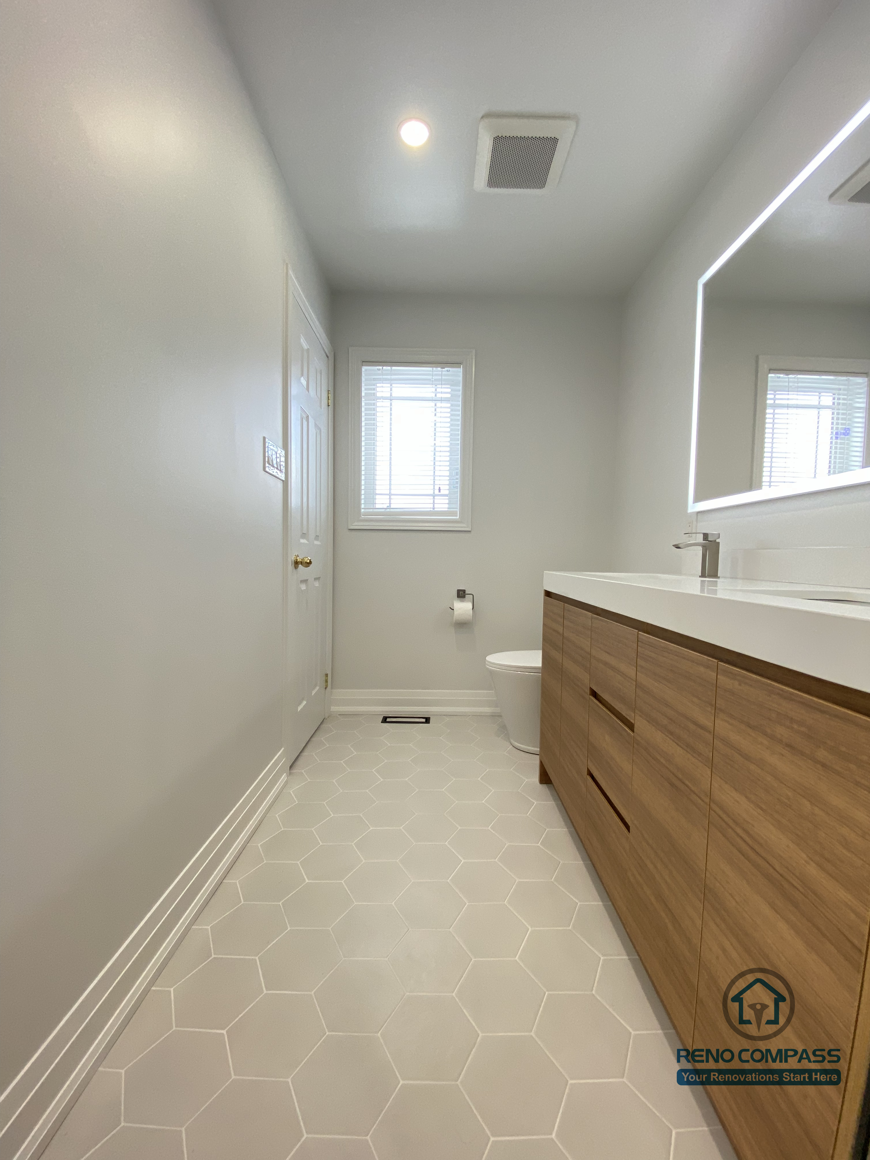This image captures the main walkway of a Markham bathroom renovation, showcasing a clean, linear layout designed for comfort and daily practicality. The hexagon tile flooring provides slip resistance and visual texture while maintaining a soft, neutral tone that keeps the space bright and calm. Recessed ceiling lighting and an integrated ventilation system ensure proper illumination and airflow, supporting long-term durability in a high-moisture environment. The wood-finish vanity on the right adds warmth and storage without crowding the circulation path. Completed by a Toronto renovation company, Reno Compass Inc., this bathroom renovation reflects thoughtful spatial planning, material coordination, and a modern, functional approach tailored for Markham homes.