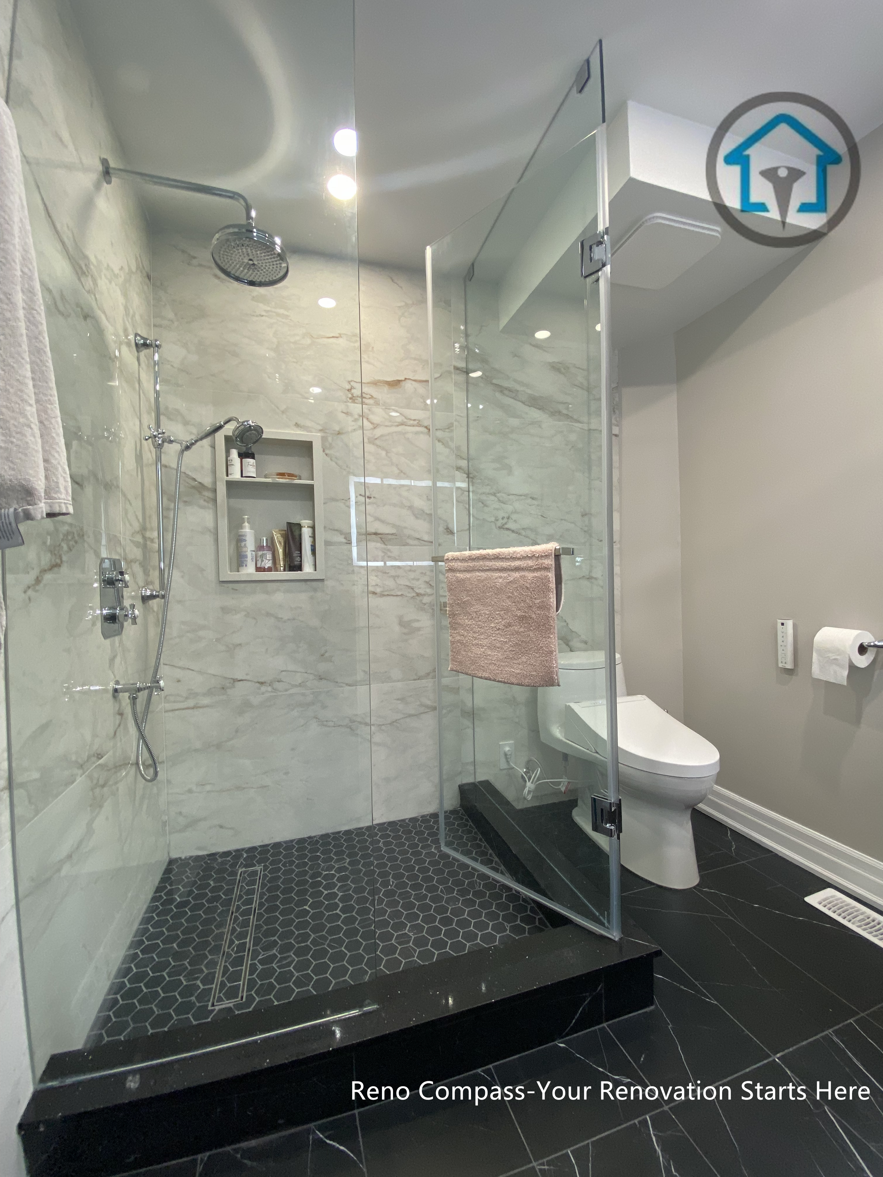 This Toronto bathroom renovation showcases a frameless glass shower paired with marble-look wall tiles and a dark hexagon mosaic shower base. The contrast between light stone surfaces and darker flooring defines the shower zone while maintaining a clean, modern appearance. Built-in shower niches provide practical storage without interrupting the visual flow, and the clear glass enclosure keeps the space feeling open and bright. The layout prioritizes comfort, durability, and ease of daily maintenance.