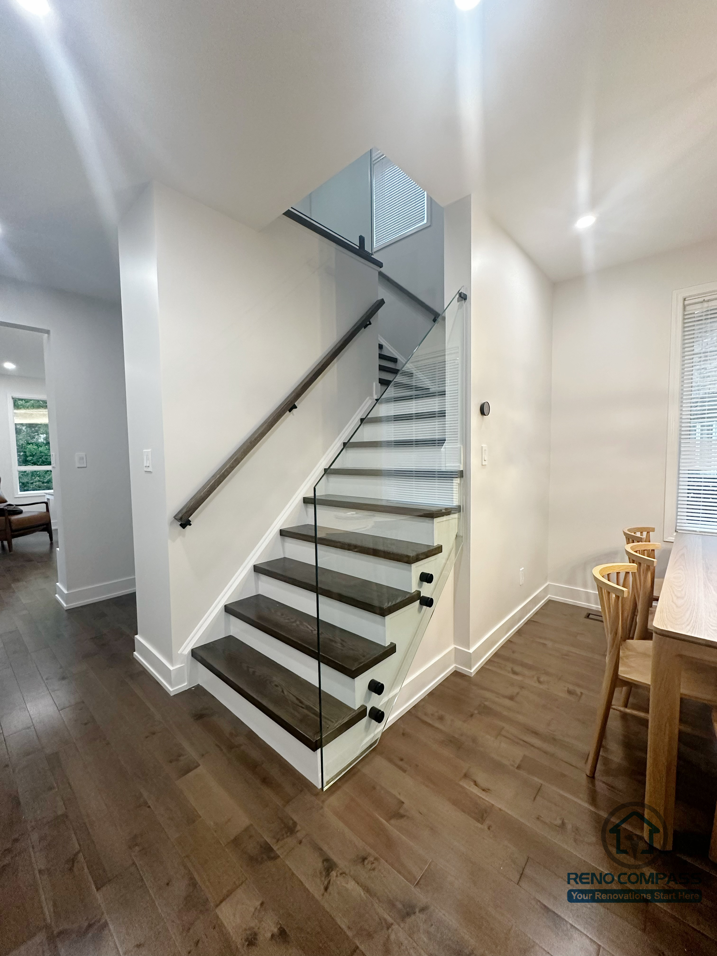 This Oakville interior renovation features a modern staircase design with solid wood treads and a full-height glass railing that enhances openness and light flow. The clean white structure is paired with warm wood flooring, creating a balanced transition between levels. The transparent railing maintains visual continuity while ensuring safety, making the staircase a functional architectural element within the open living space.