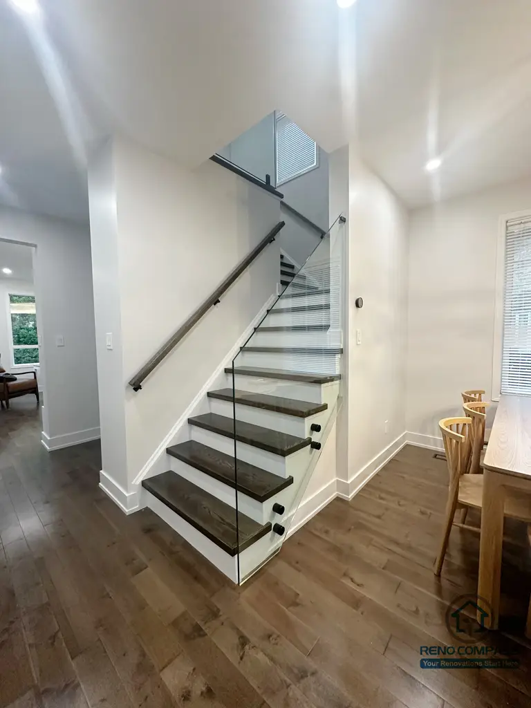This Oakville interior renovation features a modern staircase design with solid wood treads and a full-height glass railing that enhances openness and light flow. The clean white structure is paired with warm wood flooring, creating a balanced transition between levels. The transparent railing maintains visual continuity while ensuring safety, making the staircase a functional architectural element within the open living space.