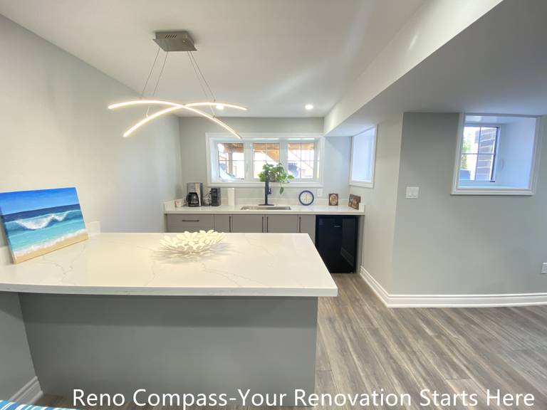 Modern basement bar renovation in Oakville featuring a large quartz island countertop, custom base cabinets, contemporary pendant lighting, vinyl plank flooring, and a compact kitchenette layout designed for entertaining and everyday use, completed by Reno Compass Inc.