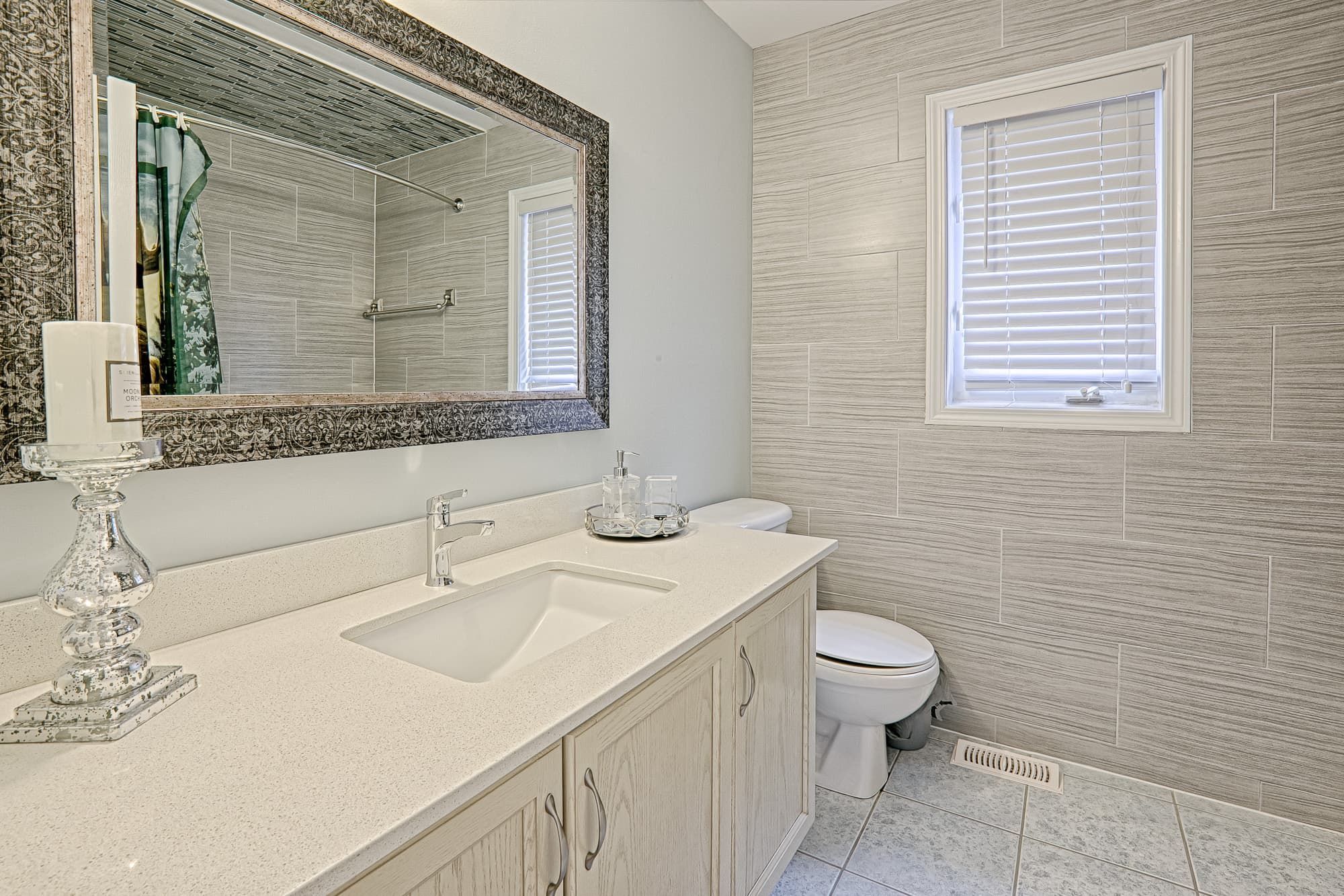 This clean bathroom vanity showcases a simple and lovely interior design with a quartz countertop, light wood cabinetry, and neutral wall tile finishes. The large framed mirror enhances brightness and visual depth, while the streamlined sink and storage provide everyday practicality. This bathroom design emphasizes cleanliness, functionality, and timeless appeal, making it suitable for family homes and long-term residential use.