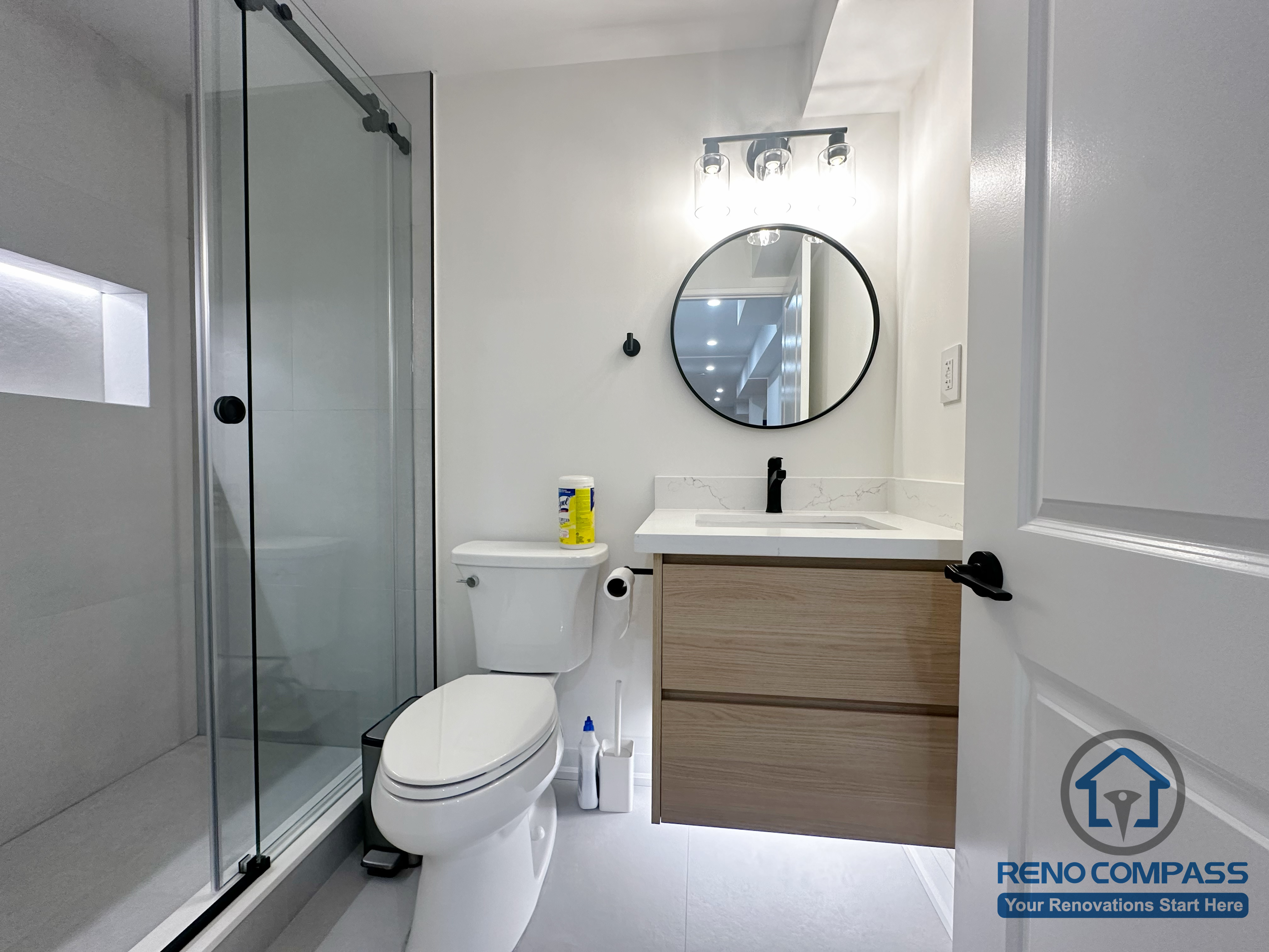 Modern basement bathroom renovation in Markham featuring a glass-enclosed shower, wall-mounted vanity with wood finish, round mirror, and clean contemporary fixtures. Designed for efficient use of space, durability, and everyday comfort.