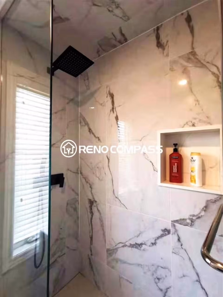 Contemporary bathroom renovation featuring a glass-enclosed shower with marble-look wall tiles, a matte black rainfall showerhead, and a built-in wall niche for convenient storage. The clean tile work and modern fixtures highlight the craftsmanship of a professional Toronto renovation company, delivering a functional, waterproof, and stylish shower space designed for everyday comfort and durability.