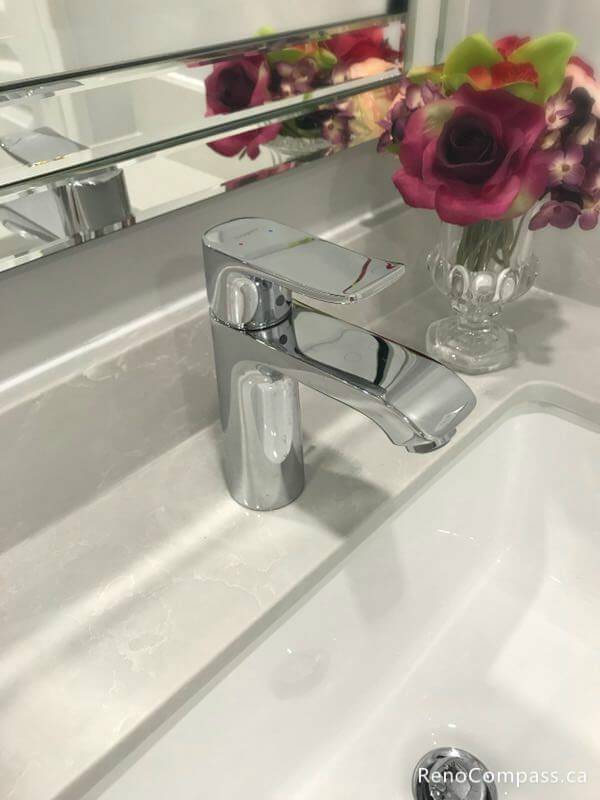 This Markham bathroom renovation detail highlights a sleek chrome faucet installed on a smooth quartz vanity countertop with an under-mount sink. The clean lines, reflective mirror finish, and decorative floral accents create a refined and modern bathroom atmosphere while complementing the overall upgraded vanity and lighting design.