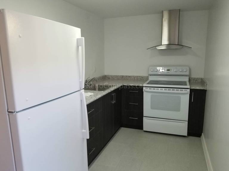 This Toronto basement renovation features a clean and functional modern kitchen designed for a basement apartment. The space includes dark wood cabinetry, quartz-style countertops, stainless steel range hood, full-size refrigerator, and durable flooring. Reno Compass Inc. optimized the layout to maximize storage and usability while maintaining a bright, minimalist look, creating a comfortable and practical basement living space for rental or family use.