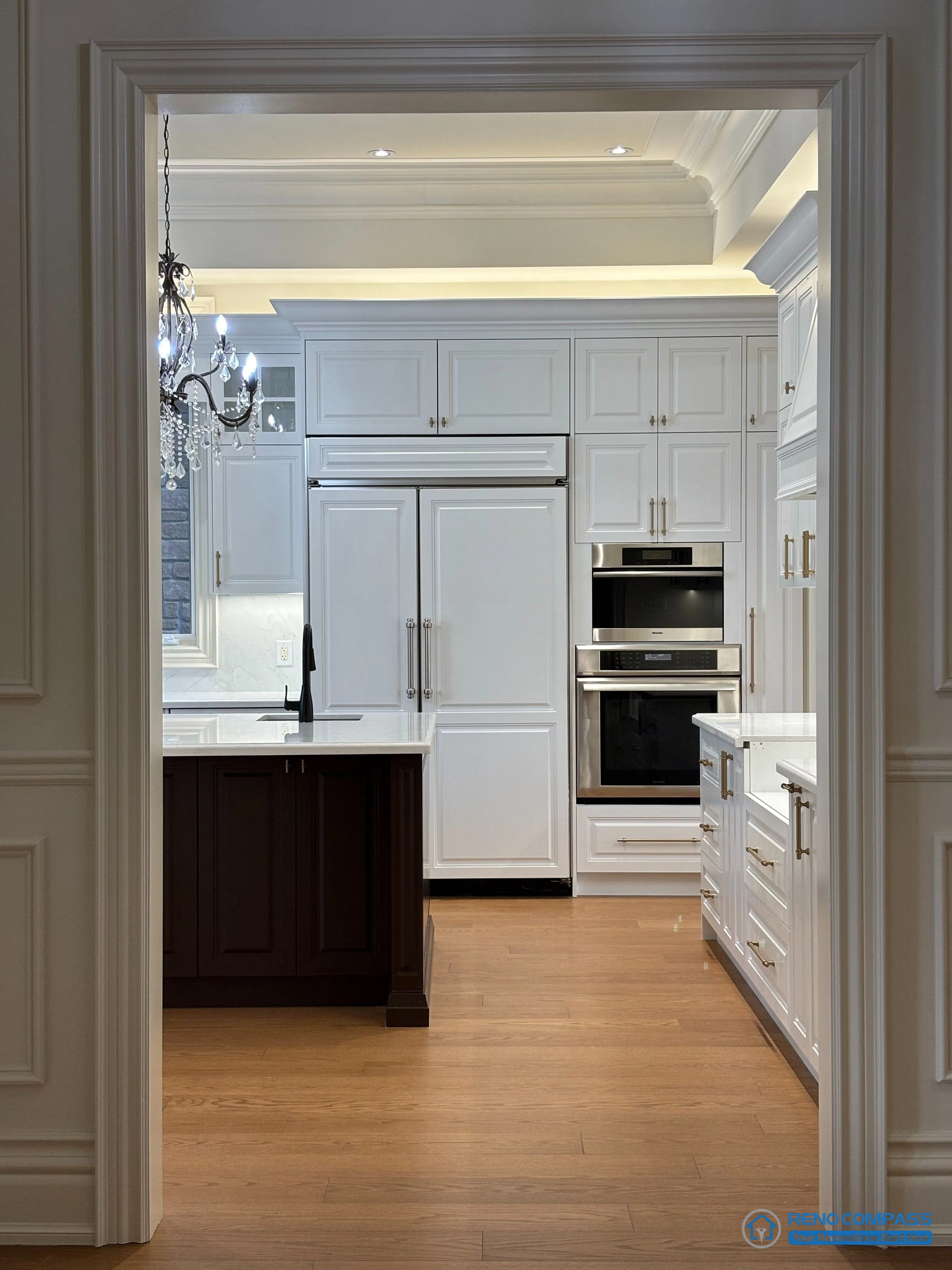 This image presents a European-style kitchen viewed through a classically detailed doorway, emphasizing architectural symmetry and proportion. Full-height cabinetry, integrated appliances, and layered ceiling moulding establish a structured and orderly kitchen composition. The contrast between the dark wood island and white cabinetry reinforces visual hierarchy, while warm hardwood flooring anchors the space. This framed perspective highlights the relationship between architectural detailing and functional kitchen design within a high-end Richmond Hill interior renovation.
