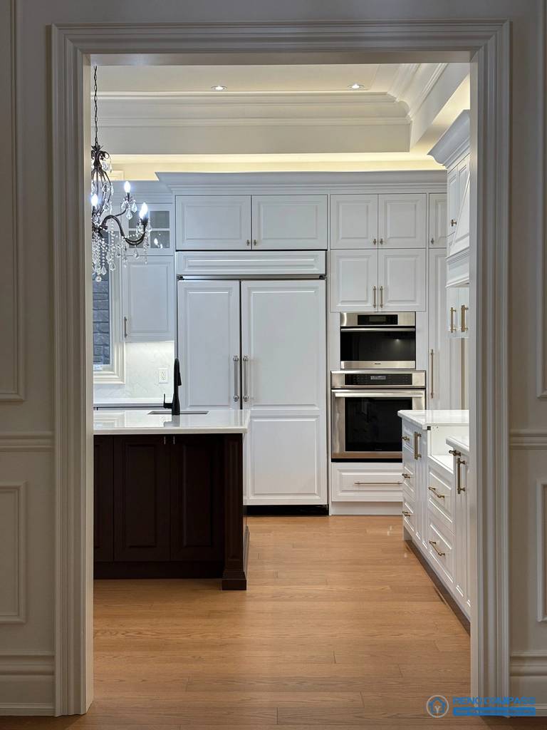 This image presents a European-style kitchen viewed through a classically detailed doorway, emphasizing architectural symmetry and proportion. Full-height cabinetry, integrated appliances, and layered ceiling moulding establish a structured and orderly kitchen composition. The contrast between the dark wood island and white cabinetry reinforces visual hierarchy, while warm hardwood flooring anchors the space. This framed perspective highlights the relationship between architectural detailing and functional kitchen design within a high-end Richmond Hill interior renovation.
