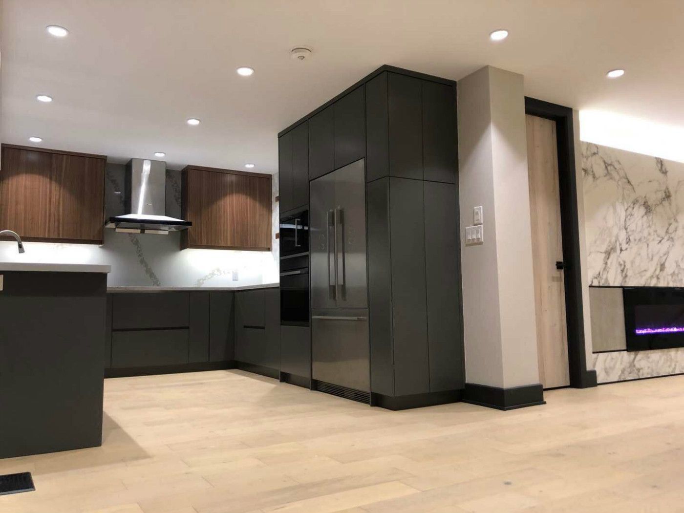 Contemporary kitchen renovation in Markham featuring custom two-tone cabinetry, built-in appliances, sleek range hood, and open-concept layout. The design integrates warm wood finishes, matte black cabinets, and modern lighting to create a functional and stylish cooking space that connects seamlessly with the living area for modern family living.
