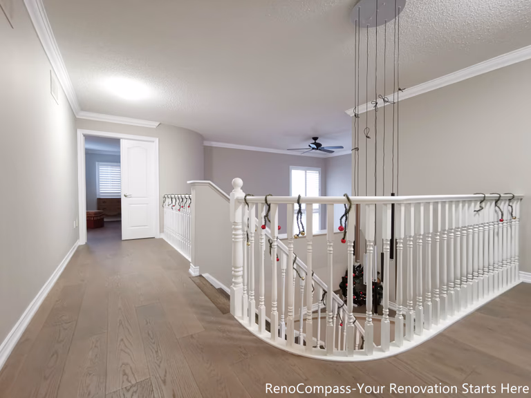 his image showcases an open hallway and staircase area completed as part of an Oakville interior renovation. Light-toned hardwood flooring extends seamlessly through the upper level, enhancing visual continuity and brightness. The curved staircase with white balusters adds architectural softness while maintaining a clean, timeless look. Neutral wall tones, crown molding, and open sightlines create a calm transition between private rooms and shared living spaces, resulting in a layout that feels both spacious and well-balanced.
